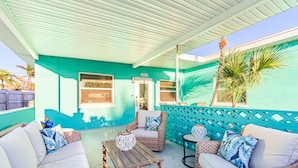 A covered patio area with wicker furniture, a wooden table, and a teal-colored wall. - Shore La Vie (Treasure Island)