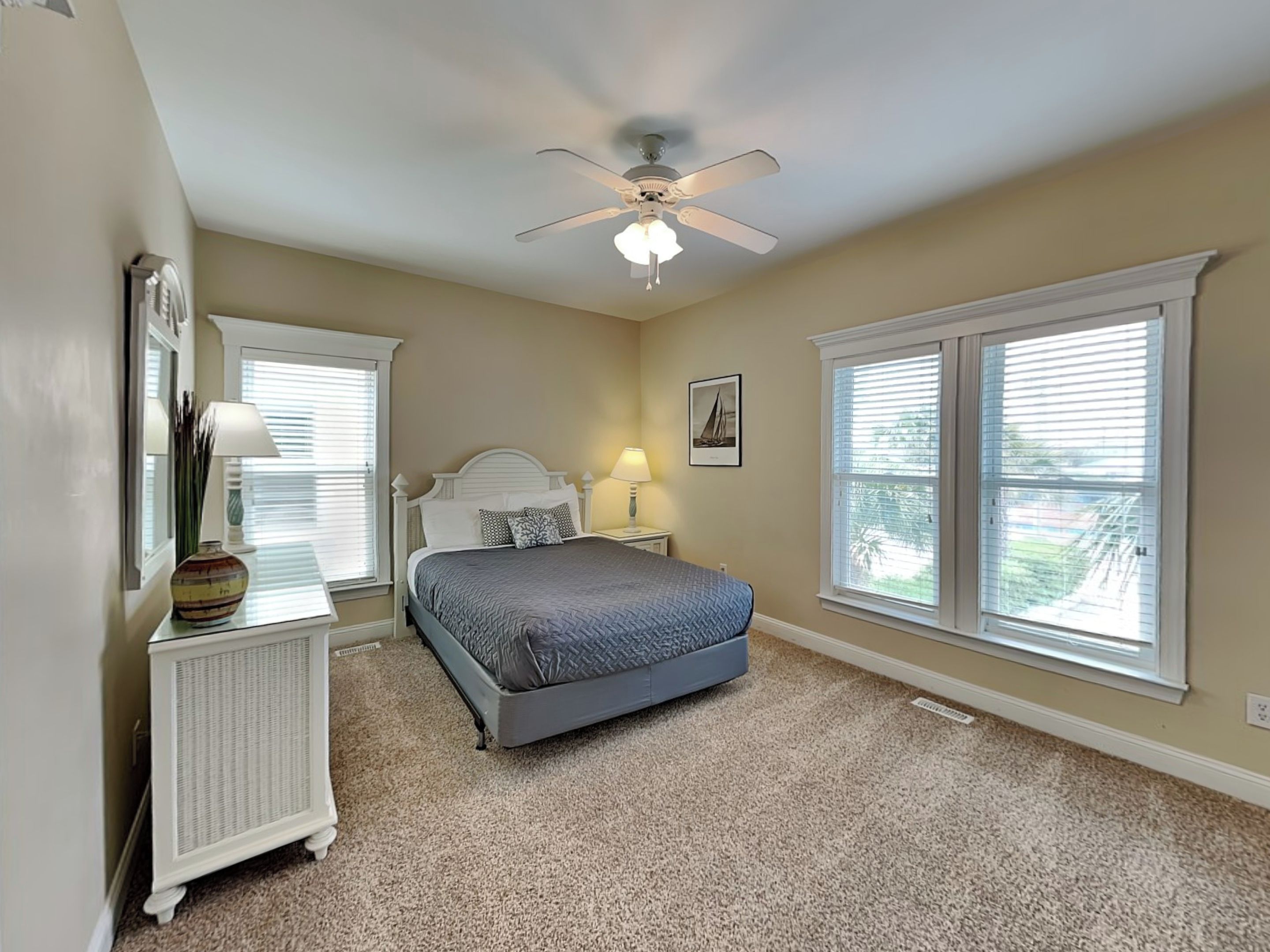 A bedroom with a bed, a ceiling fan, two windows with blinds, a nightstand with a lamp, and a dresser.
