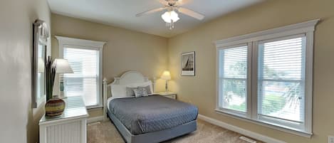 A bedroom with a bed, a ceiling fan, two windows with blinds, a nightstand with a lamp, and a dresser.