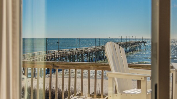 A beach view with a wooden pier extending into the ocean, a white beach chair, and a wooden deck. - Jolly Dave's by Topsail Realty (Holly Ridge)