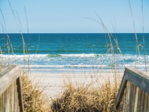 A beach view with waves, a wooden walkway, and tall grass in the foreground. - Dock Holliday by Topsail Realty (Holly Ridge)