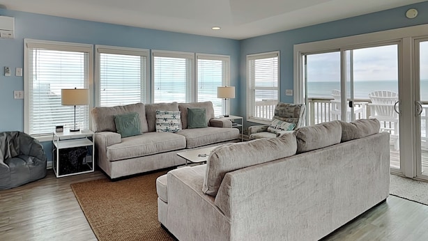 A modern living room with a large sectional sofa, a bean bag chair, and a coffee table. There are multiple windows with blinds, a lamp on a side table, and a view of the ocean through the glass doors. - Luna Escape by Topsail Realty (Holly Ridge)