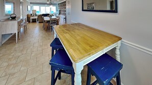 Eine Küche mit einem Holztisch und blauen Barhocker, ein Essbereich mit einer Couch und Sesseln, sowie ein Blick ins Freie durch große Fenster.
