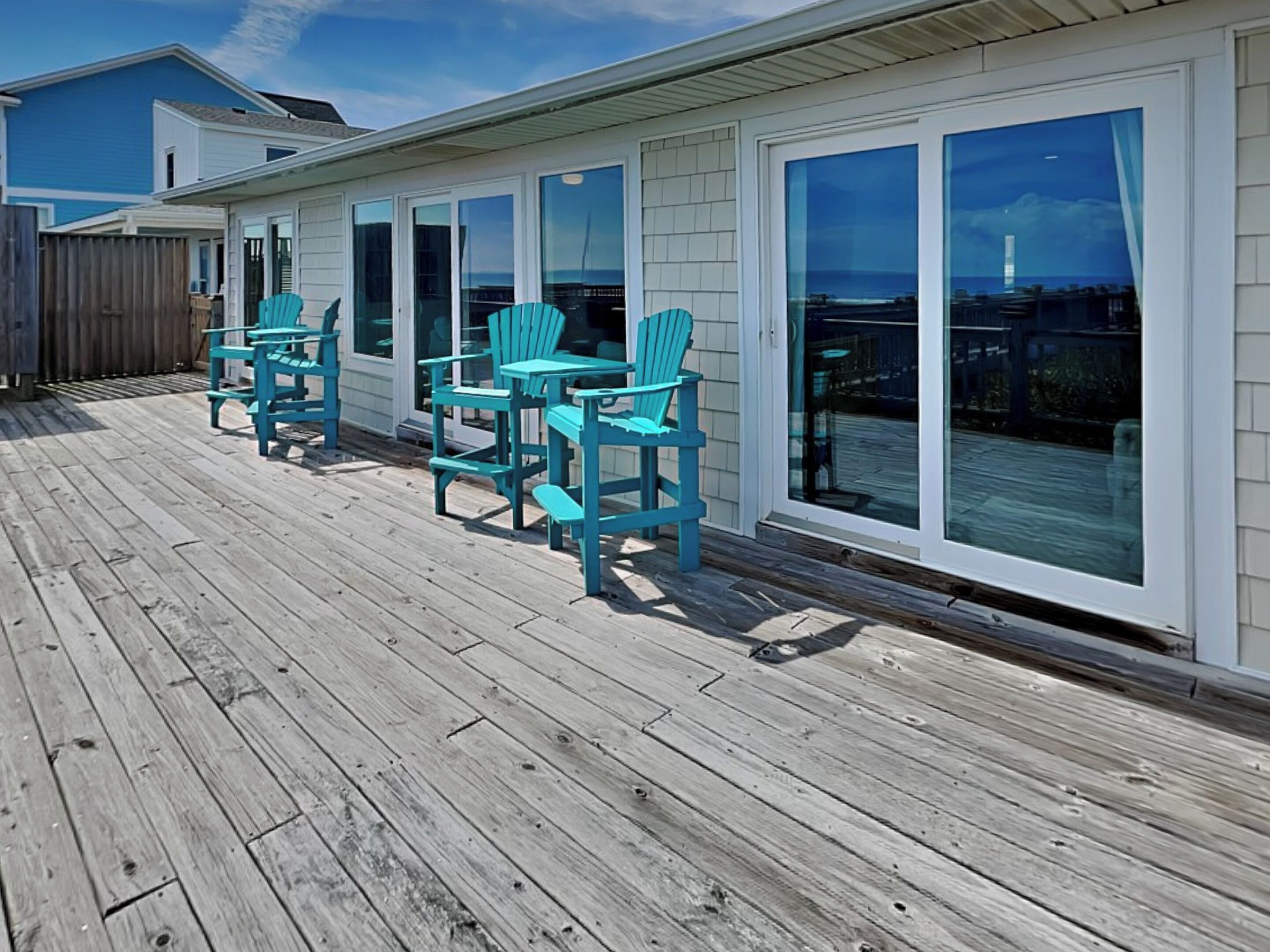 A wooden deck with turquoise chairs and a table, large sliding glass doors, and a view of the ocean.