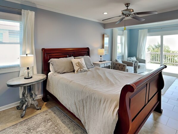 A bedroom with a wooden bed, two bedside tables with lamps, a ceiling fan, and a view of the outdoors. - Tournesol by Topsail Realty (Holly Ridge)