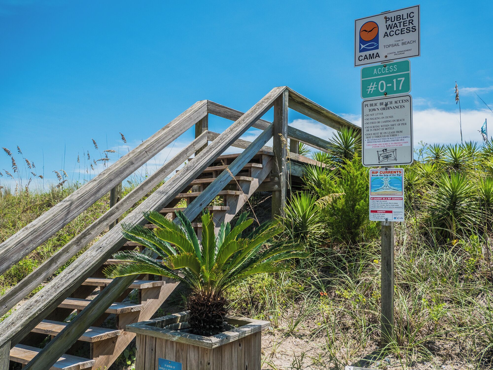 Una scala in legno conduce a una spiaggia, con segnali che indicano l'accesso pubblico all'acqua e avvisi di sicurezza.