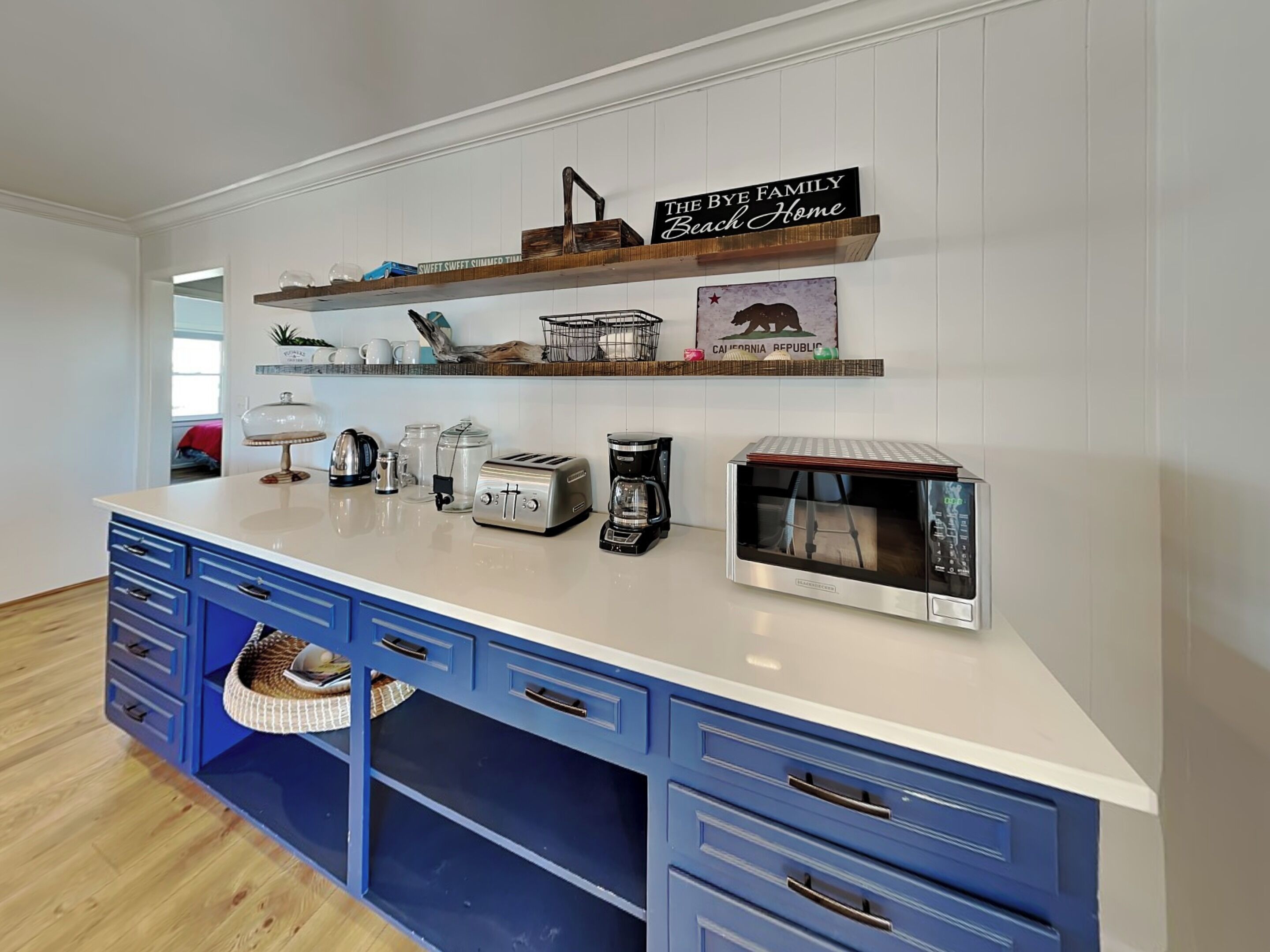 A kitchen with a white countertop, blue cabinets, and a coffee maker. There are shelves above the counter with decorative items and a sign that reads "The Bye Family Beach Home."
