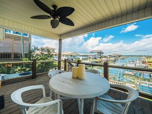 A covered outdoor deck with a round table and white chairs, overlooking a marina with boats and houses.