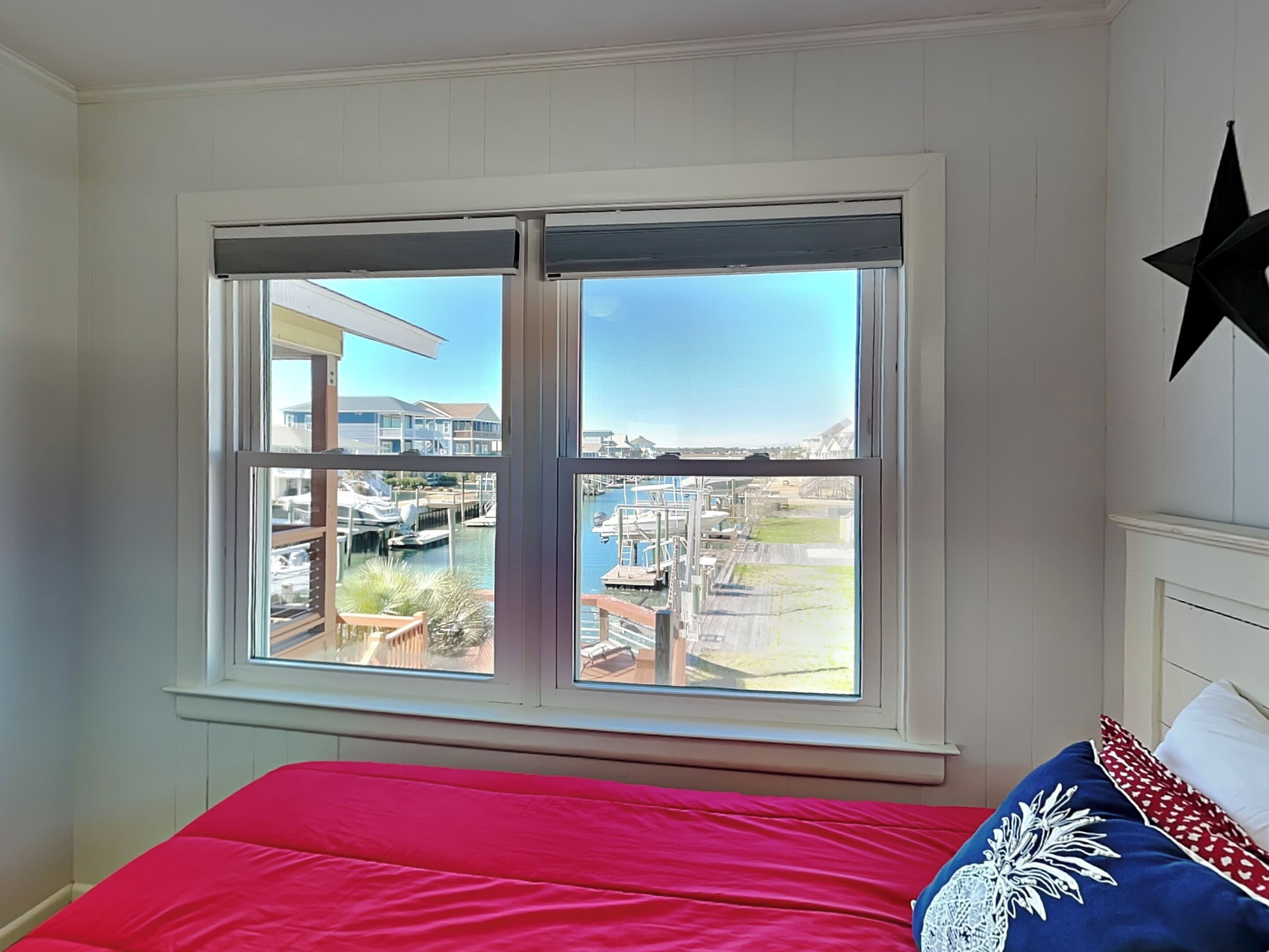 Ein Schlafzimmer mit großem Fenster mit Blick auf eine Marina, einem Bett mit roter Bettwäsche und einer Stern-Dekoration an der Wand.