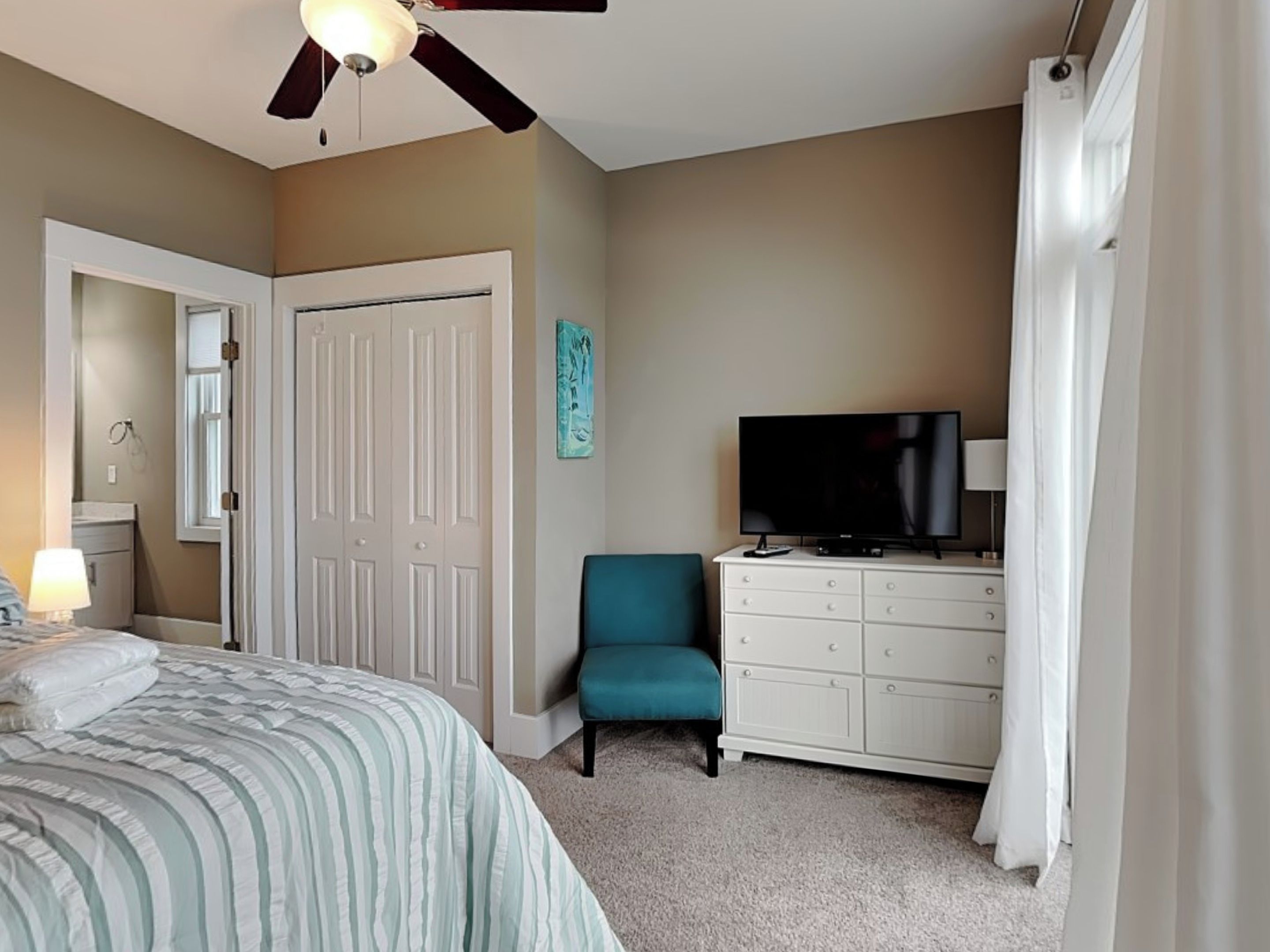Une chambre à coucher avec un lit, un téléviseur posé sur une commode, une chaise et un ventilateur de plafond.