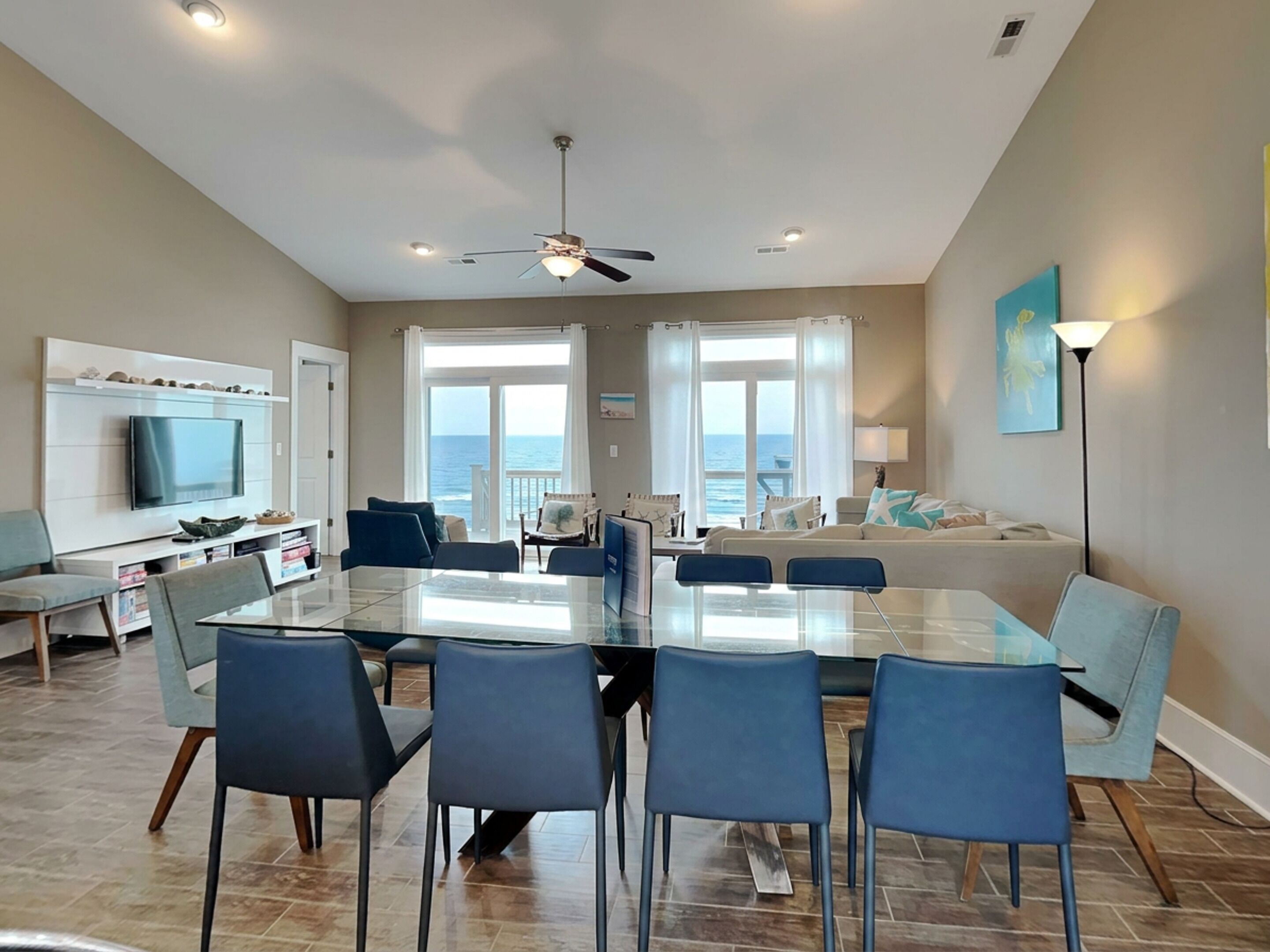 A modern living room with a glass dining table, blue chairs, a TV, and a view of the ocean.