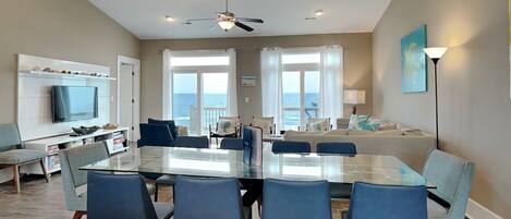 A modern living room with a glass dining table, blue chairs, a TV, and a view of the ocean.