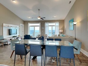 Uma sala de estar moderna com uma mesa de jantar de vidro, cadeiras azuis, uma televisão e vista para o oceano.