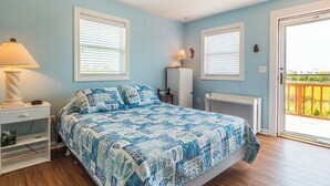 A bedroom with a bed, two windows with blinds, a nightstand with a lamp, a dresser, and a door leading to a balcony. - Sunset Bay by Topsail Realty (Sneads Ferry)