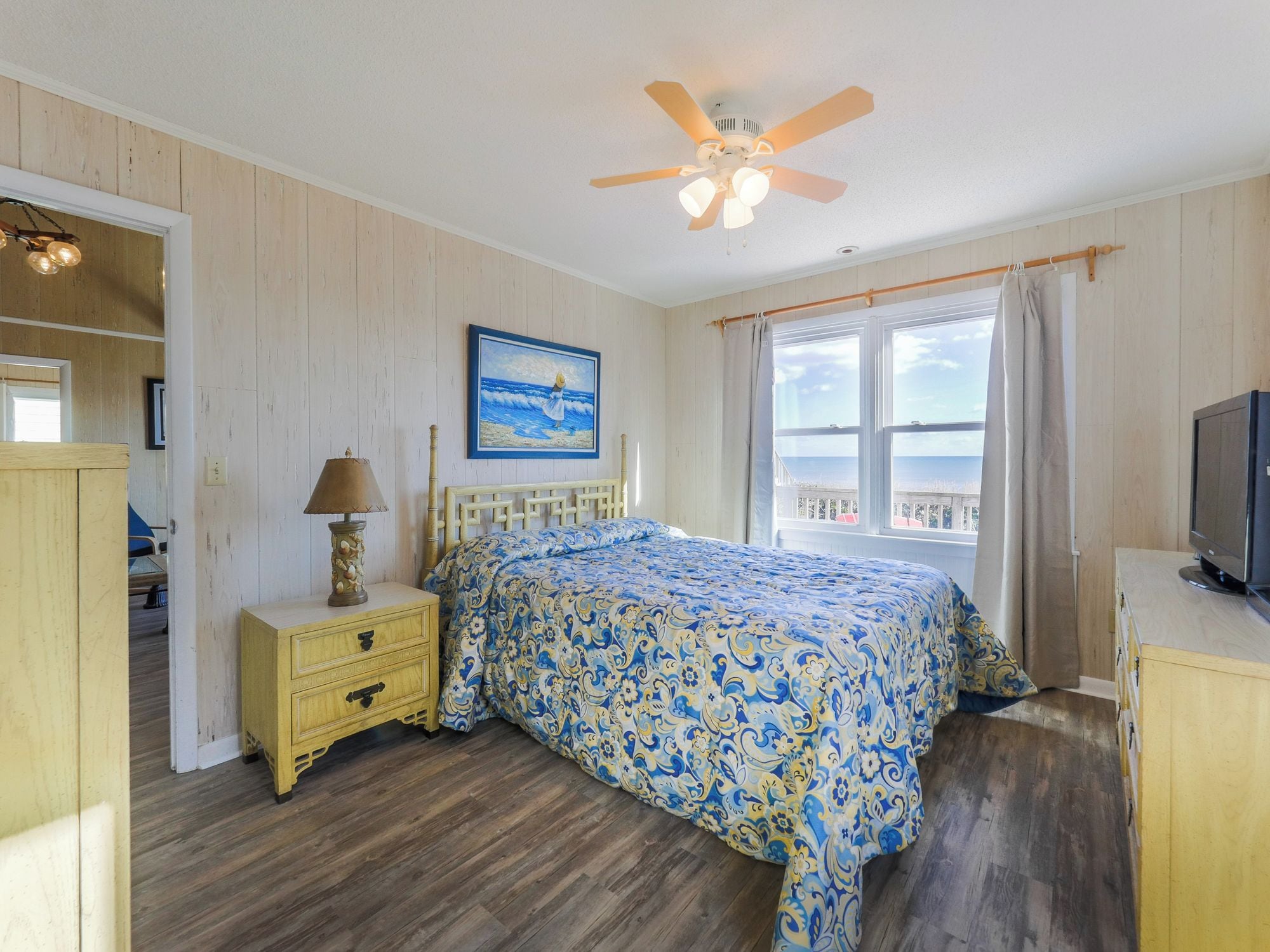 A bedroom with a bed, a nightstand, a ceiling fan, a window with a view, and a TV.