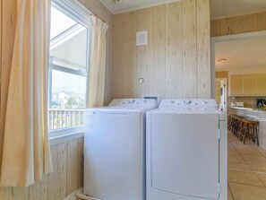 A laundry room with two white washing machines, wooden walls, a window with curtains, and a kitchen area in the background.