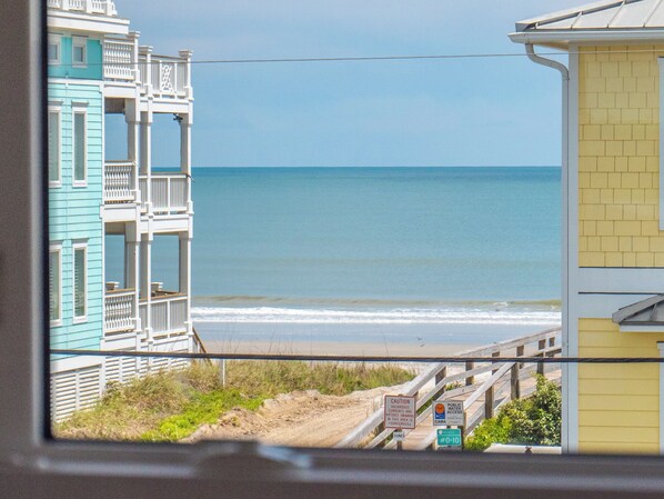 A view of a beach with a wooden boardwalk, a sign indicating public water access, and a building with a yellow facade.