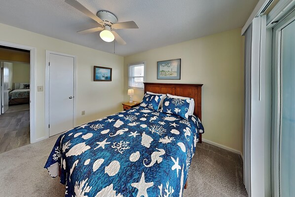 A bedroom with a bed, a ceiling fan, a nightstand, two framed pictures, and a door leading to another room.