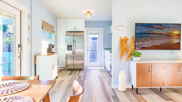 A modern kitchen with white cabinets, a stainless steel refrigerator, and a dining area with a wooden table and chairs. - Se La Vie (Treasure Island)