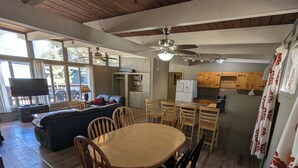 Un ampio soggiorno con un divano grande, un tavolo da pranzo in legno e sedie. Si vede anche una televisione, una ventola a soffitto e un angolo cottura sullo sfondo.
