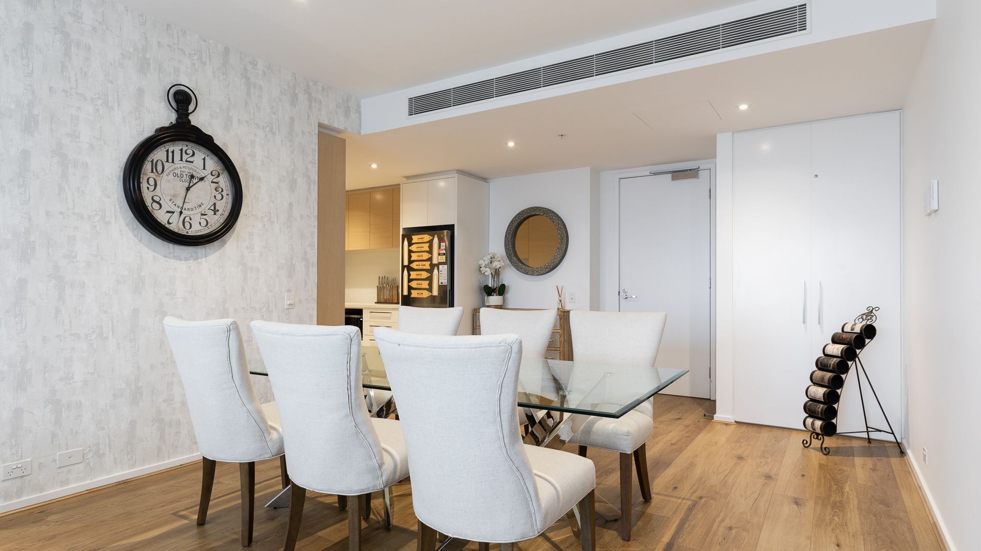 A dining area with a glass table and four chairs, a wall clock, a mirror, and a wine rack.