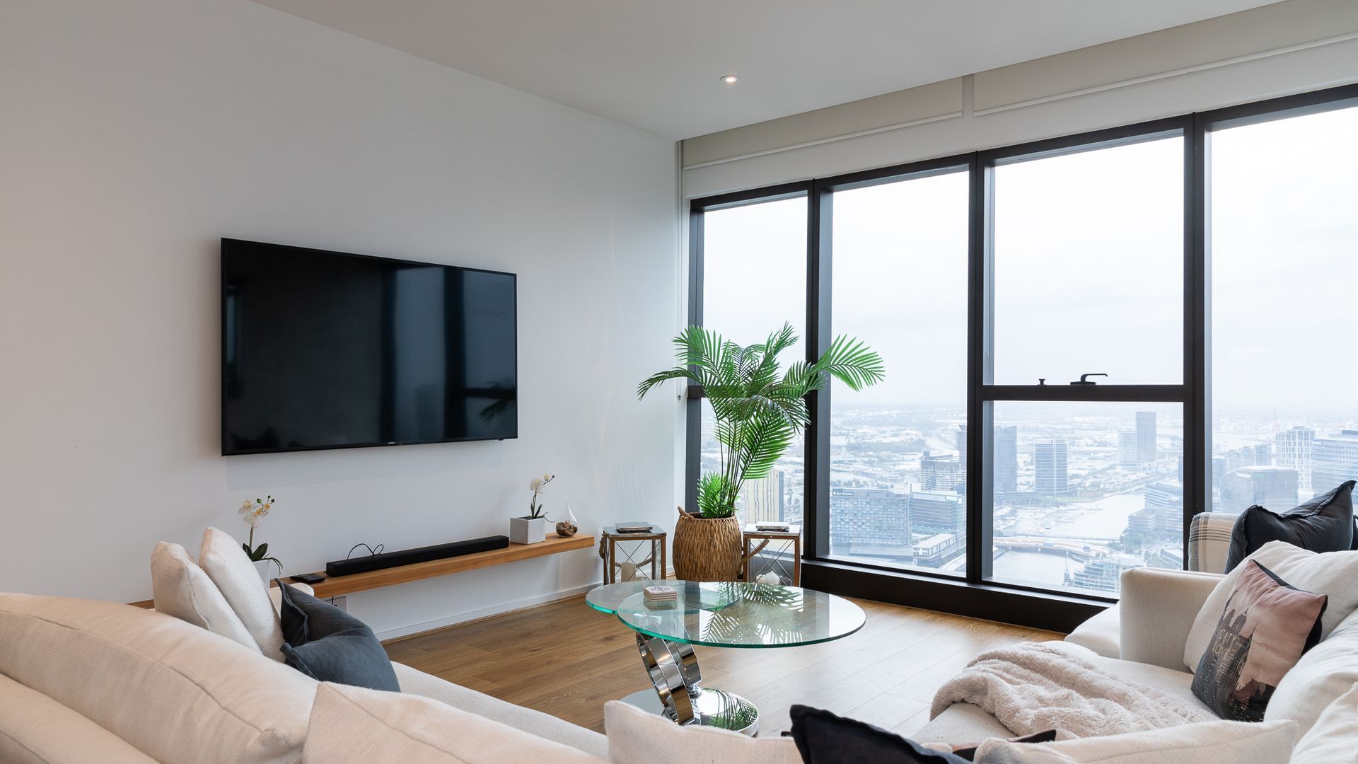 A modern living room with a large TV, a glass coffee table, and a view of the city through large windows.