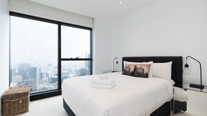 A modern hotel room with a large window offering a city view, a neatly made bed with white linens, and a small side table with a lamp.