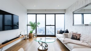 A modern living room with a large window, a flat-screen TV, a glass coffee table, and a sofa with cushions.