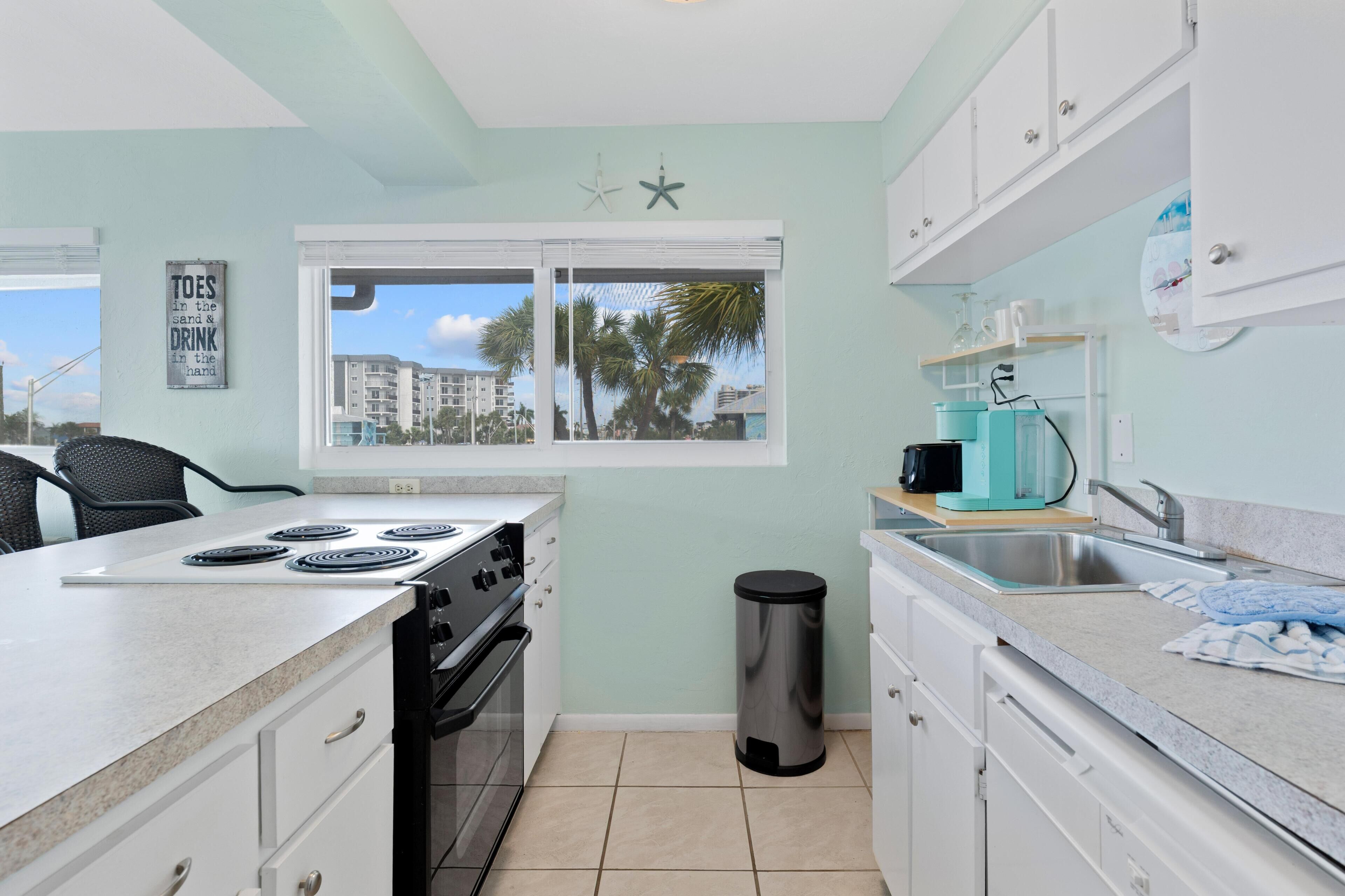 A kitchen with a stove, sink, and window overlooking palm trees.