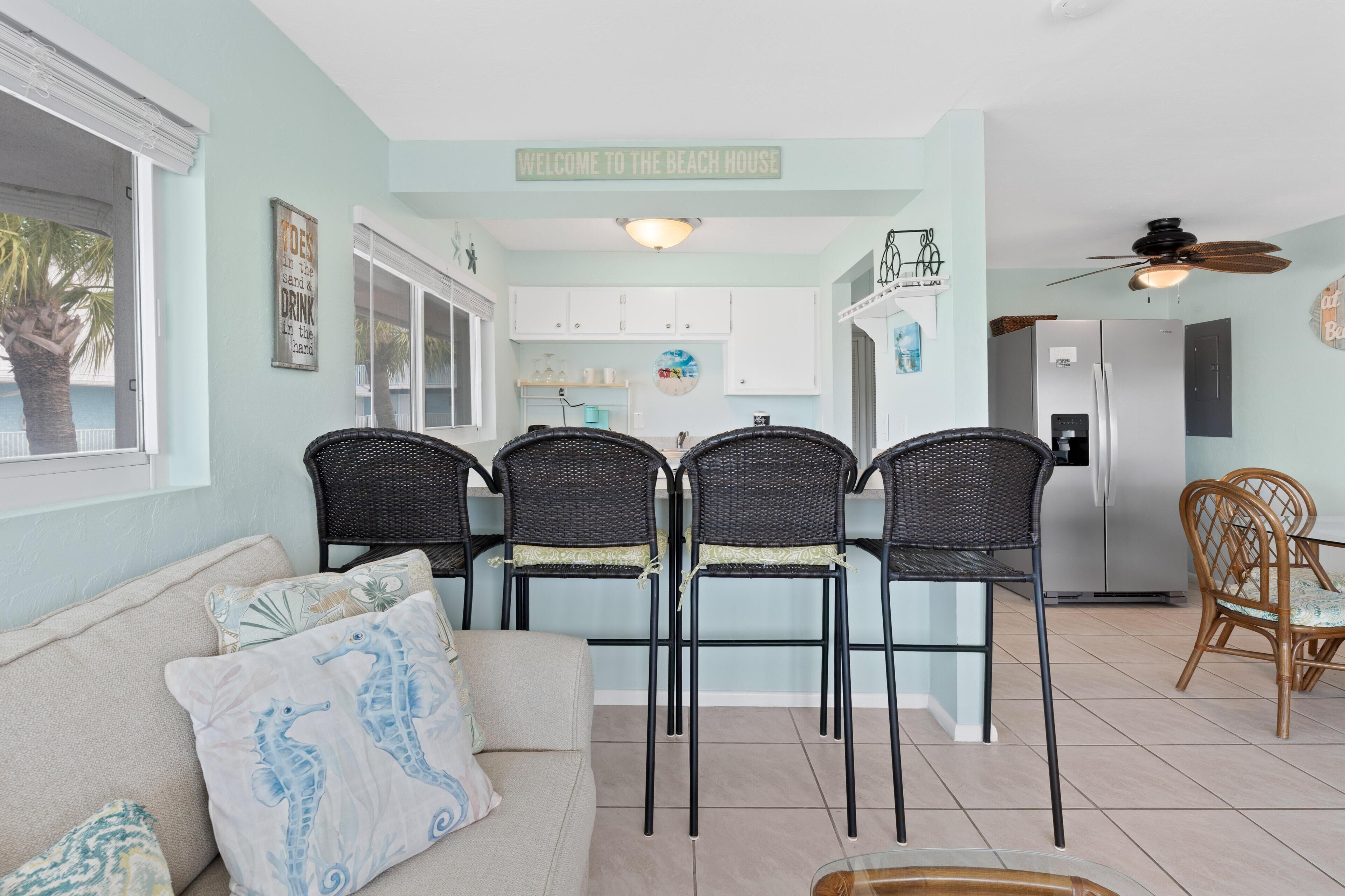 Eine Küche mit einem Schild, auf dem 'Welcome to the Beach House' steht, ein Essbereich mit Korbstühlen, ein Kühlschrank und ein Deckenventilator.