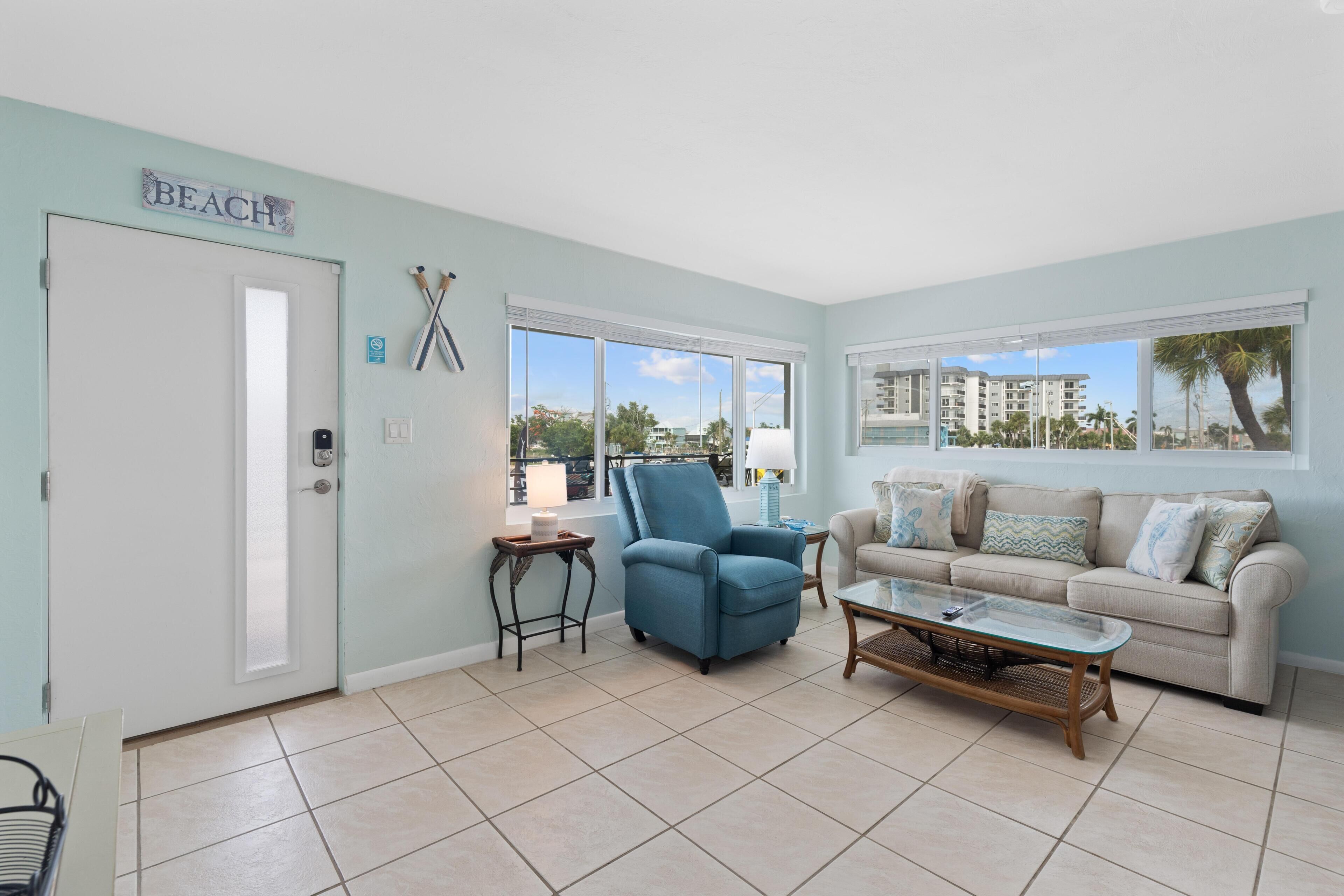 A living room with a blue armchair, beige sofa, and a coffee table. A 'BEACH' sign is on the wall, and there are large windows with a view of buildings and palm trees.