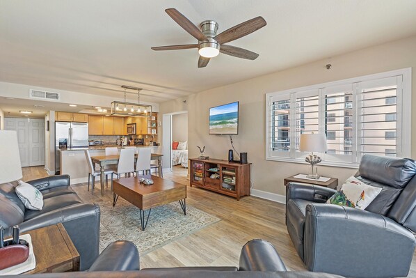 A spacious living area with a dining table, leather sofas, a ceiling fan, and a kitchen in the background.