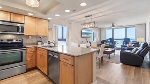 A modern kitchen with granite countertops, wooden cabinets, and stainless steel appliances. There is a dining area with a gray sofa and a view of the ocean through large windows. - Sunrise 302 (New Smyrna Beach)