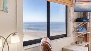 A room with a large window overlooking a beach, a television, and a shelf with various items. - Sunrise 502 (New Smyrna Beach)