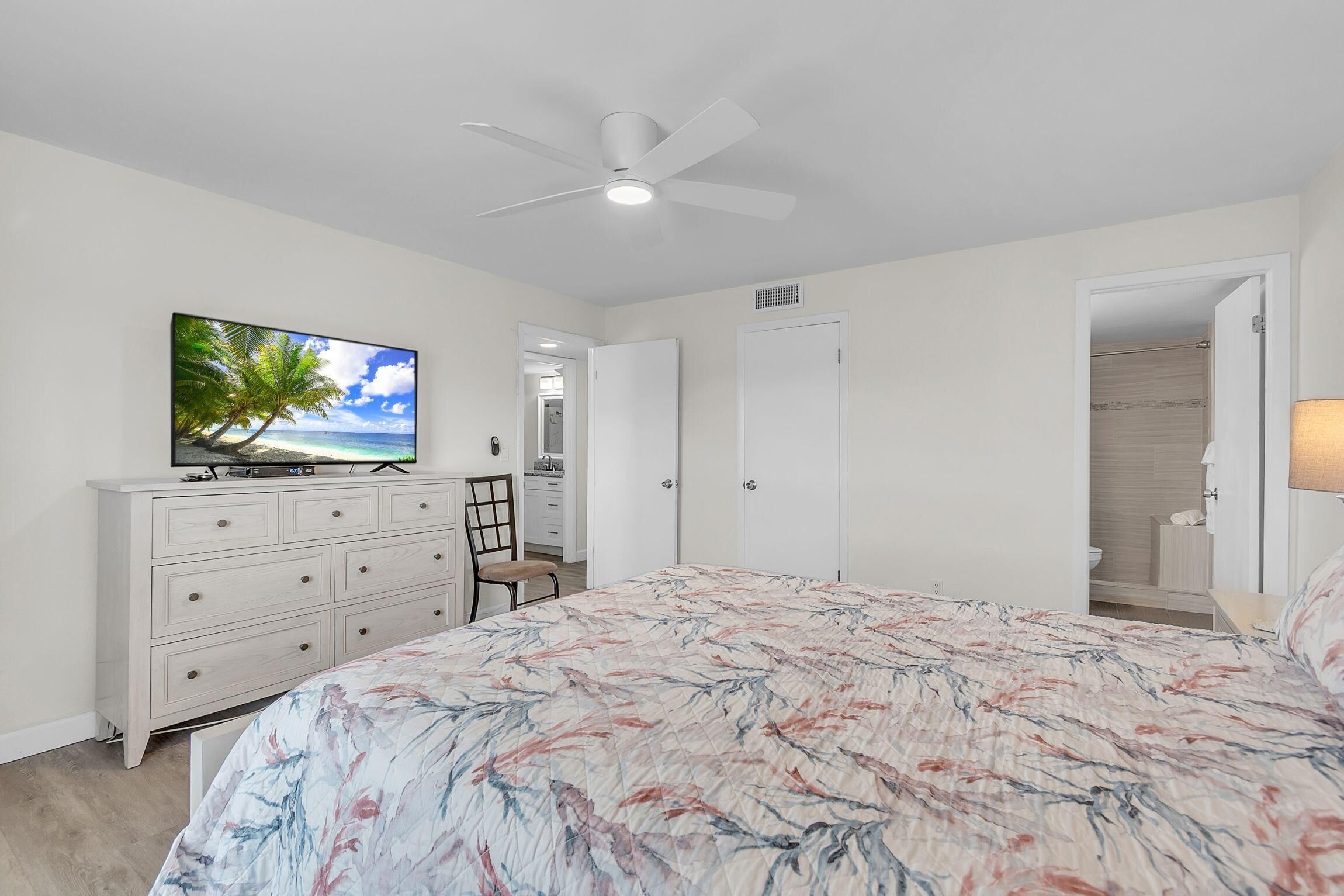 A bedroom with a bed, a TV on a dresser, a ceiling fan, and a door leading to a bathroom.