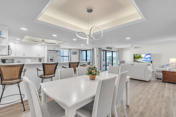 A modern kitchen with white cabinets, a dining area with a white table and chairs, and a living room with a sofa and a TV.