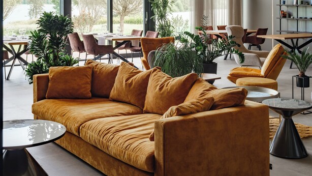 Un vaste espace de vie comprenant un grand canapé, des tables et des chaises, de grandes fenêtres et des plantes d’intérieur.