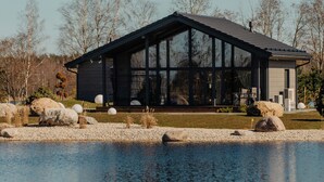 Une maison moderne dotée d’une grande façade en verre, située au bord d’un plan d’eau, et entourée de rochers et d’arbres.