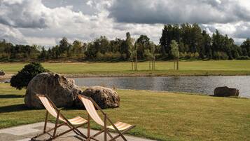 Un paisaje tranquilo junto al lago, con dos sillas de playa a rayas sobre una plataforma de concreto, rodeado de césped y un lago en calma, bajo un cielo parcialmente nublado.