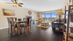 A spacious living room with a dining area, a ceiling fan, and a large window offering a view of the city skyline. - Sea Club #102 - Another Day in Paradise (Indian Shores)