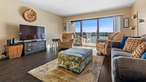 A living room with a flat-screen TV, a sofa, a loveseat, a coffee table, and a large window offering a view of the outdoors. - Sea Club #102 - Another Day in Paradise (Indian Shores)