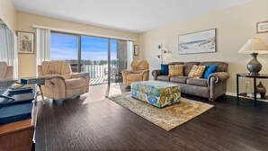 A spacious living room with a large window, a sofa set, a coffee table, and a desk with a computer. - Sea Club #102 - Another Day in Paradise (Indian Shores)