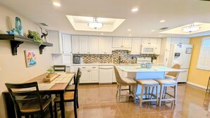 A modern kitchen with white cabinets, a central island with bar stools, and a dining area with a wooden table and chairs. - Sea Club #29 - Our Sunrise Nest (Indian Shores)