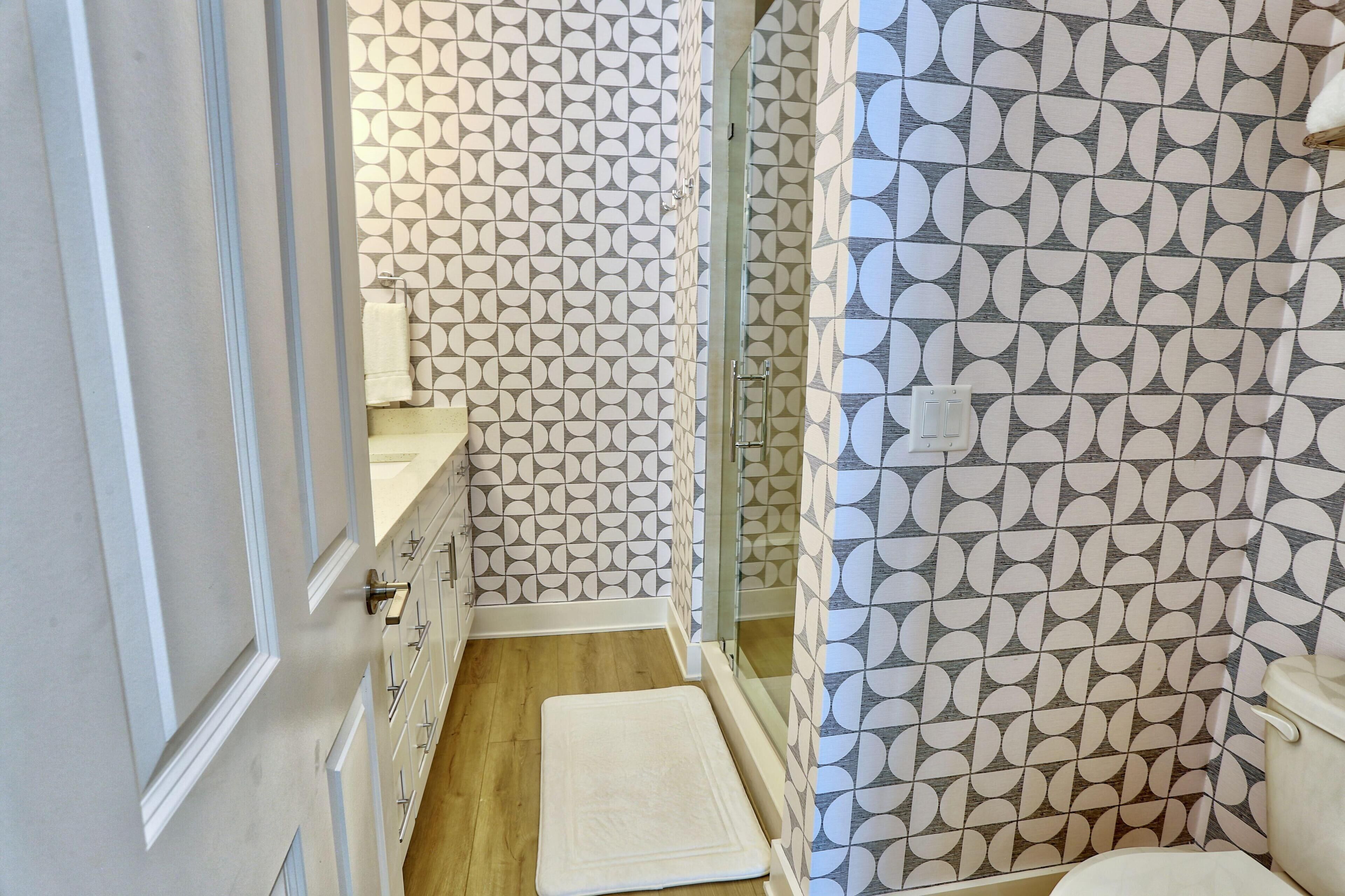 Un baño estrecho con paredes con diseño, suelo de madera, una toalla blanca y una mampara de vidrio.