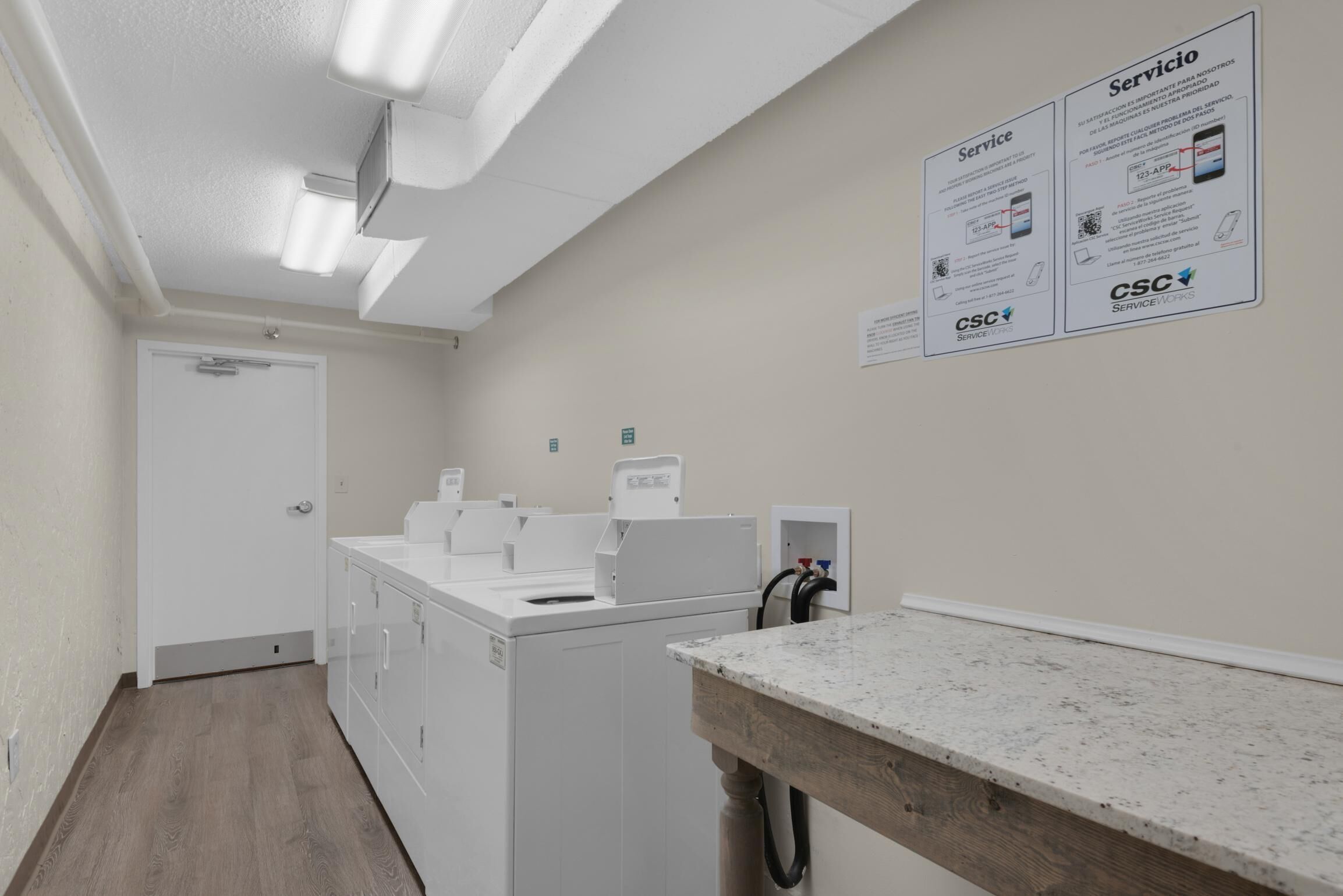 Une buanderie équipée d’un lave-linge, d’un sèche-linge et d’un plan de travail avec un évier. Des panneaux sont affichés sur le mur pour faire la publicité de services de blanchisserie.