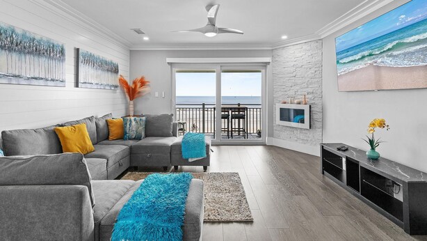 Een moderne woonkamer met een grijze modulaire bank, een grote flatscreen televisie en een balkon met uitzicht op de oceaan.
