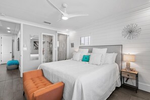 Une chambre avec un grand lit, un ventilateur de plafond, une table de chevet avec une lampe et une petite chaise orange.