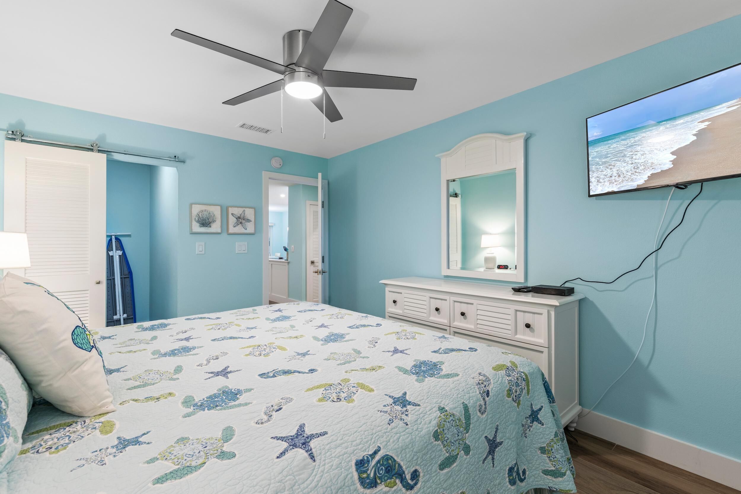 A bedroom with a bed, a ceiling fan, a dresser, a television, and a door leading to another room.