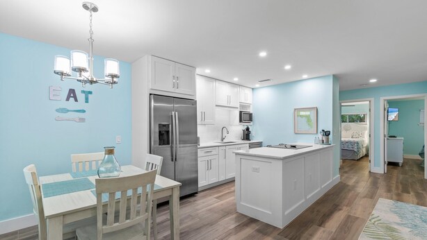 A modern kitchen with white cabinetry, a stainless steel refrigerator, and a dining area with a wooden table and chairs. - Coronado Arms Condominiums B-18 (New Smyrna Beach)