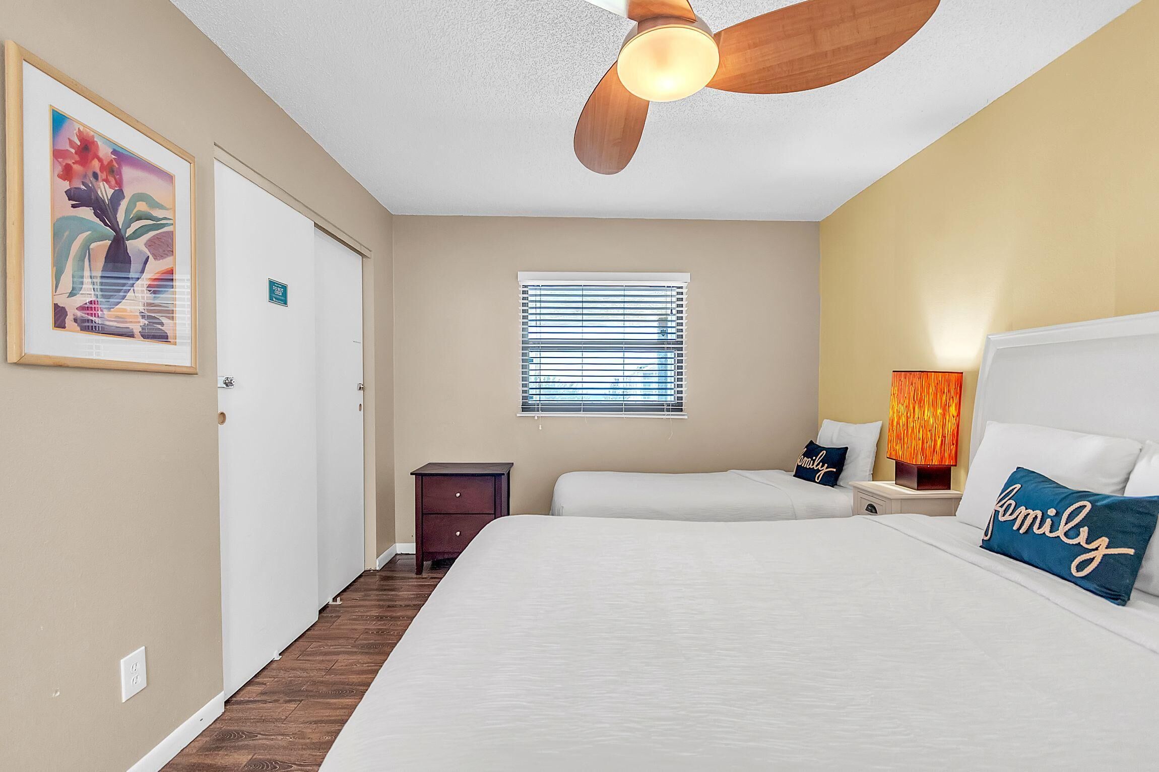A hotel room with a bed, a nightstand, a lamp, a ceiling fan, a window with blinds, and a framed picture on the wall.